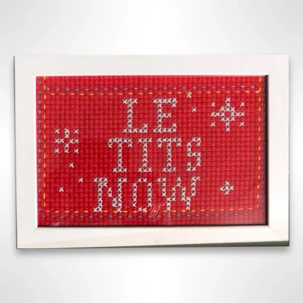 Håndlaget broderi med teksten "Le Tits Now" på rødt Aida-stoff. Morsom dekorasjon med hvit ramme, størrelse 10x15 cm. Perfekt som gave eller veggpynt.