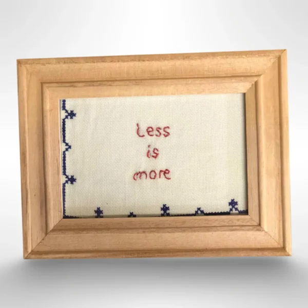 Håndlaget broderi med teksten "Less is more" på beige Aida-stoff, dekorert med blå bord. Leveres med en ramme i naturlig tre. Perfekt som gave eller veggpynt.